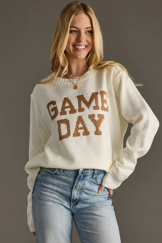 Woman wearing a cream sweater with 'GAME DAY' text and blue jeans against a gray background