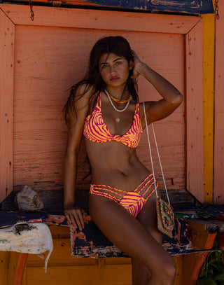 The model is wearing a vibrant bikini set featuring:A bold pink and orange zebra-print bikini top, designed with a knot detail at the center, offering a playful and eye-catching look.A matching bikini bottom with a high-leg cut and triple-strap design on the sides, adding a stylish and modern touch while enhancing the silhouette.
Layered gold necklaces, including a delicate pendant necklace and a beaded choker
front view